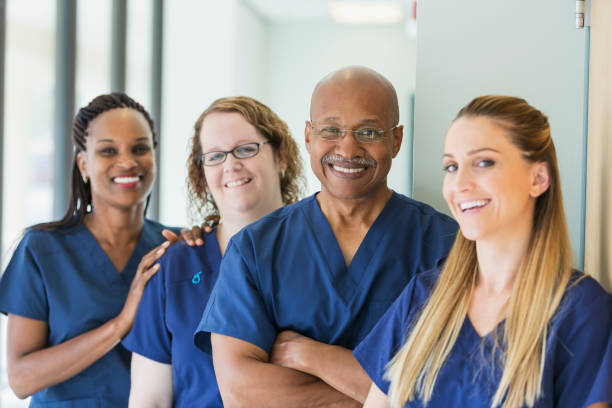 A team of four multi-ethnic medical professionals standing in a corridor, wearing blue scrubs, smiling at the camera. The focus is on the African-American senior man, in his 60s.